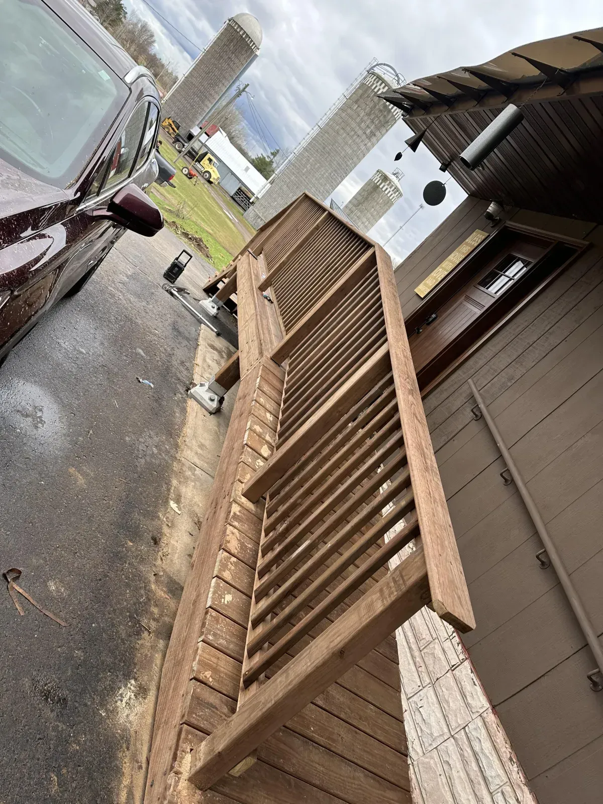 Wet driveway beside a brown house and wooden deck, with a maroon car parked on the left.