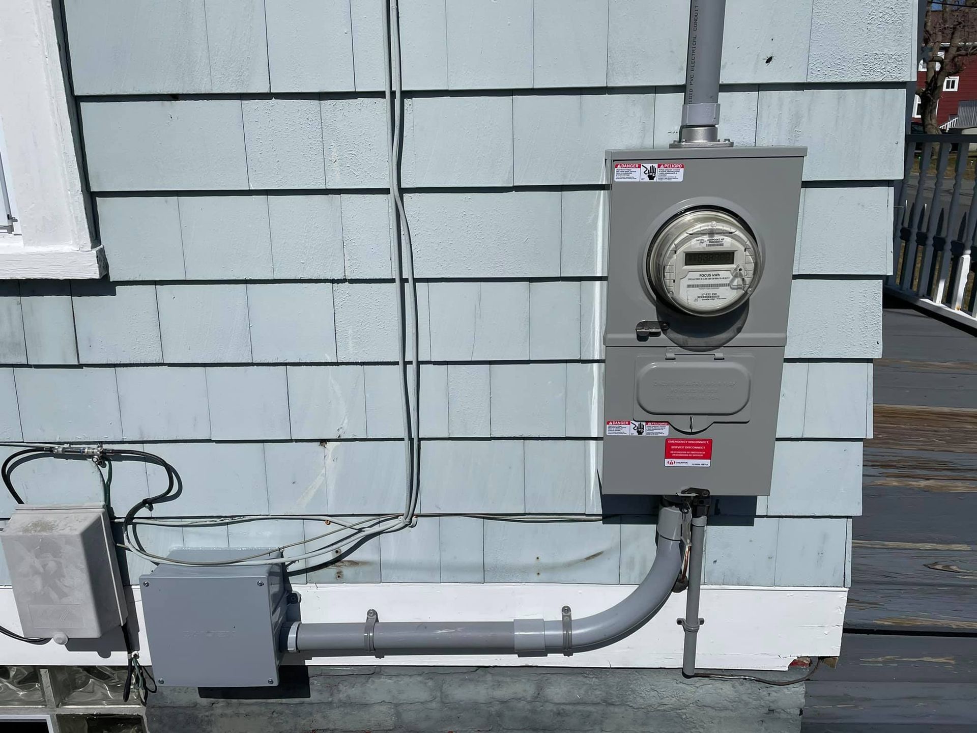 Gray electrical meter box on a blue-gray house exterior. Conduit runs to a smaller box on the left.
