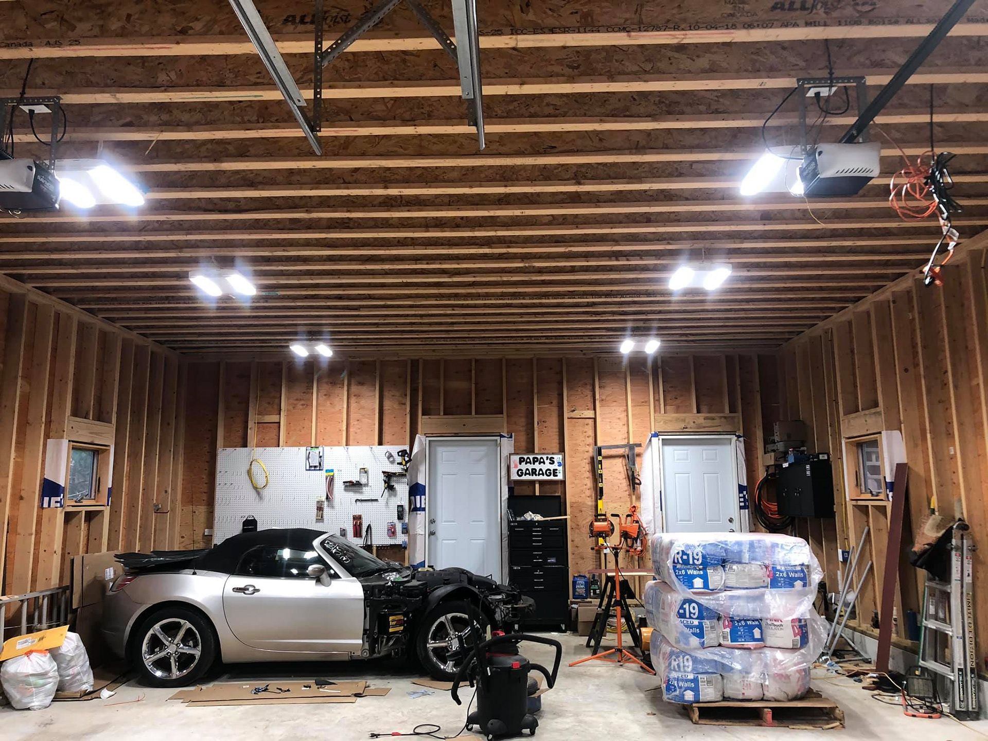 Silver convertible sports car in an unfinished garage with tools and supplies.