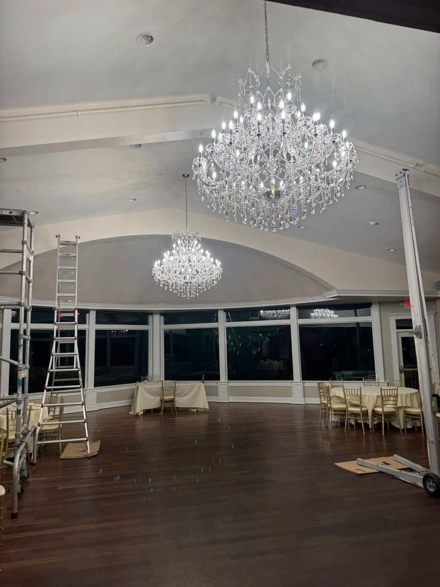 Two large crystal chandeliers hang in a brightly lit event space with dark wood floors and large windows.