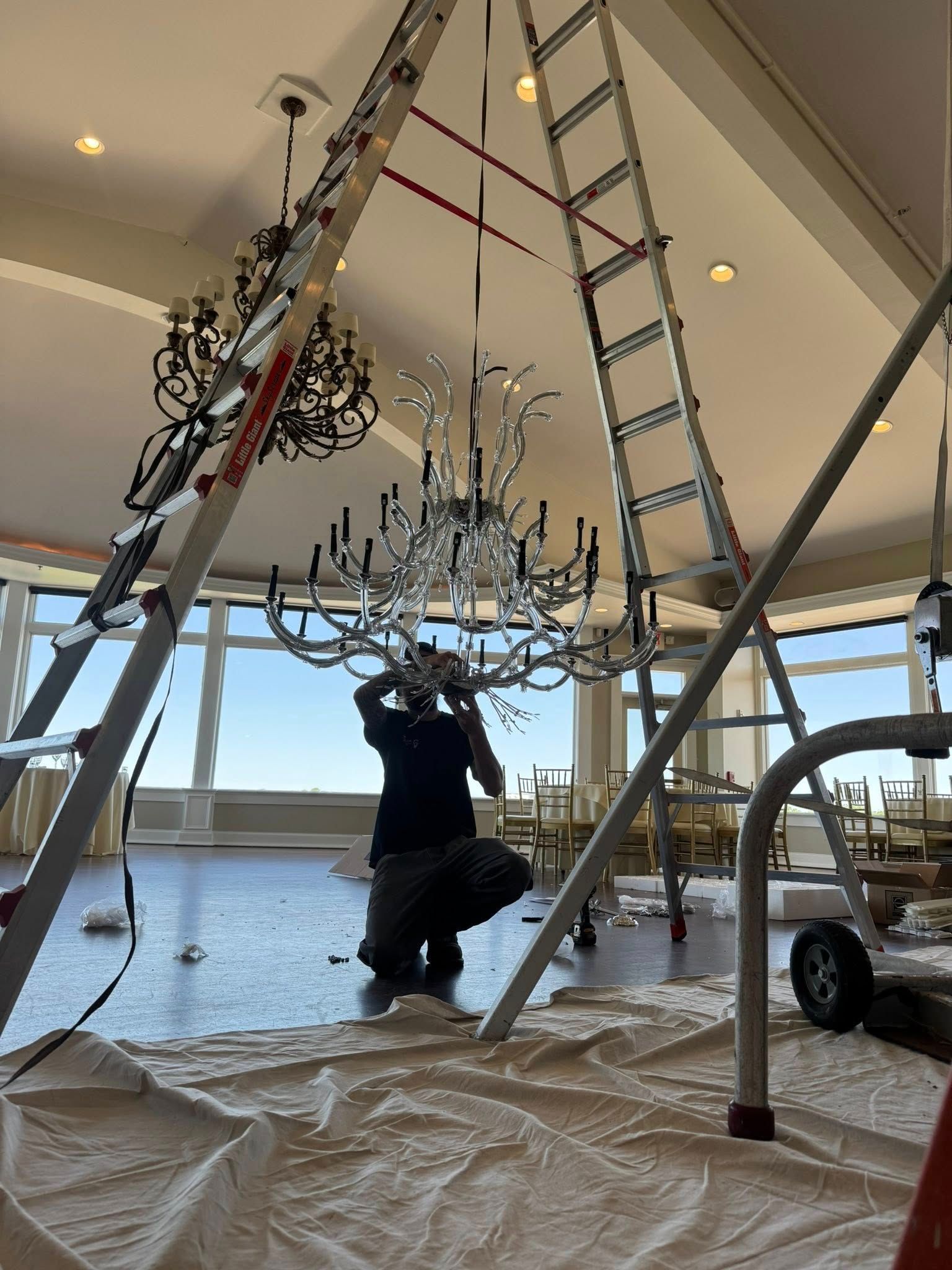 Person installing a chandelier, standing on a ladder in a room with a large window.