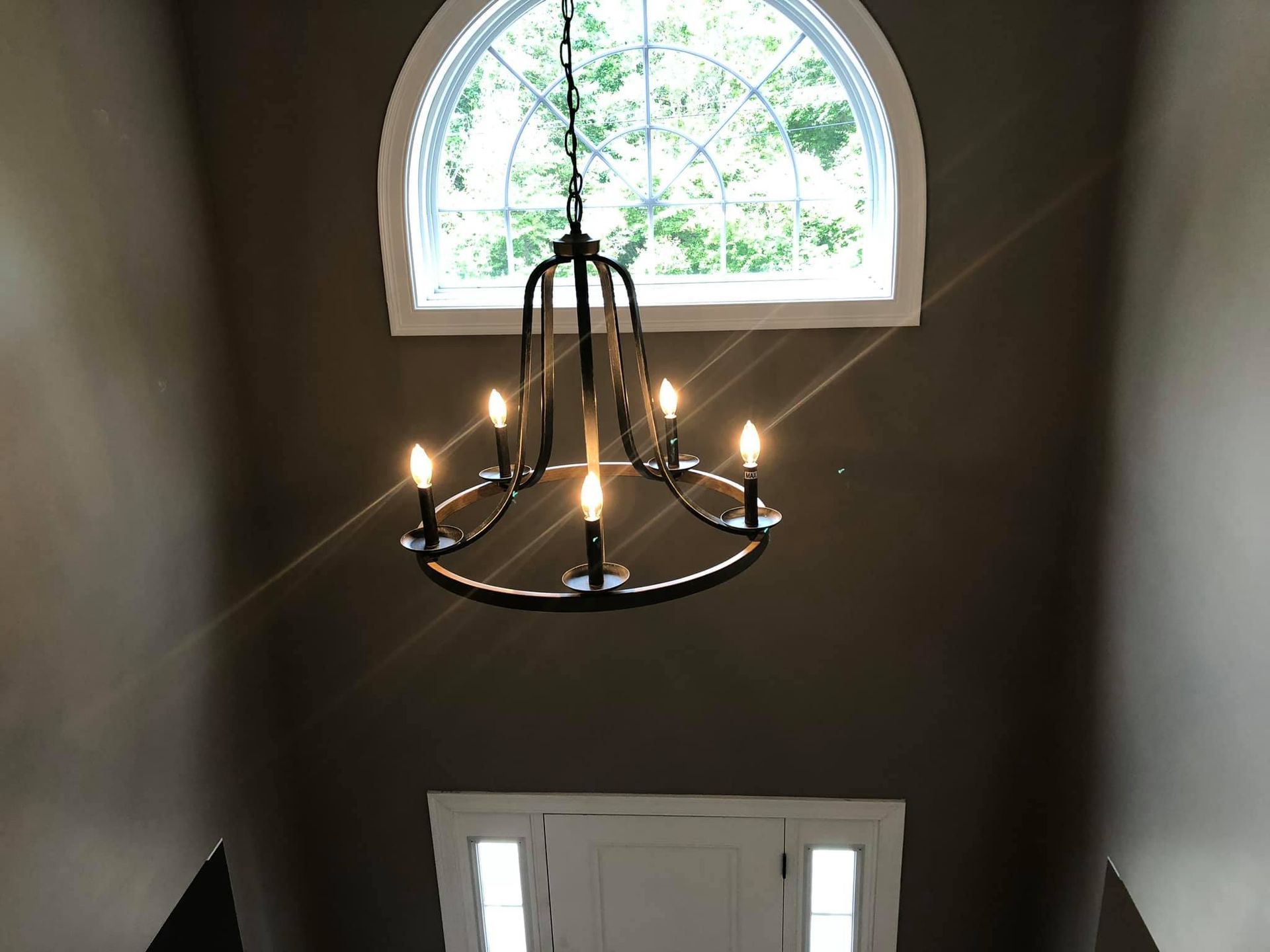 Chandelier hangs in a stairwell with a large arched window above a white door. The walls are painted a dark brown.