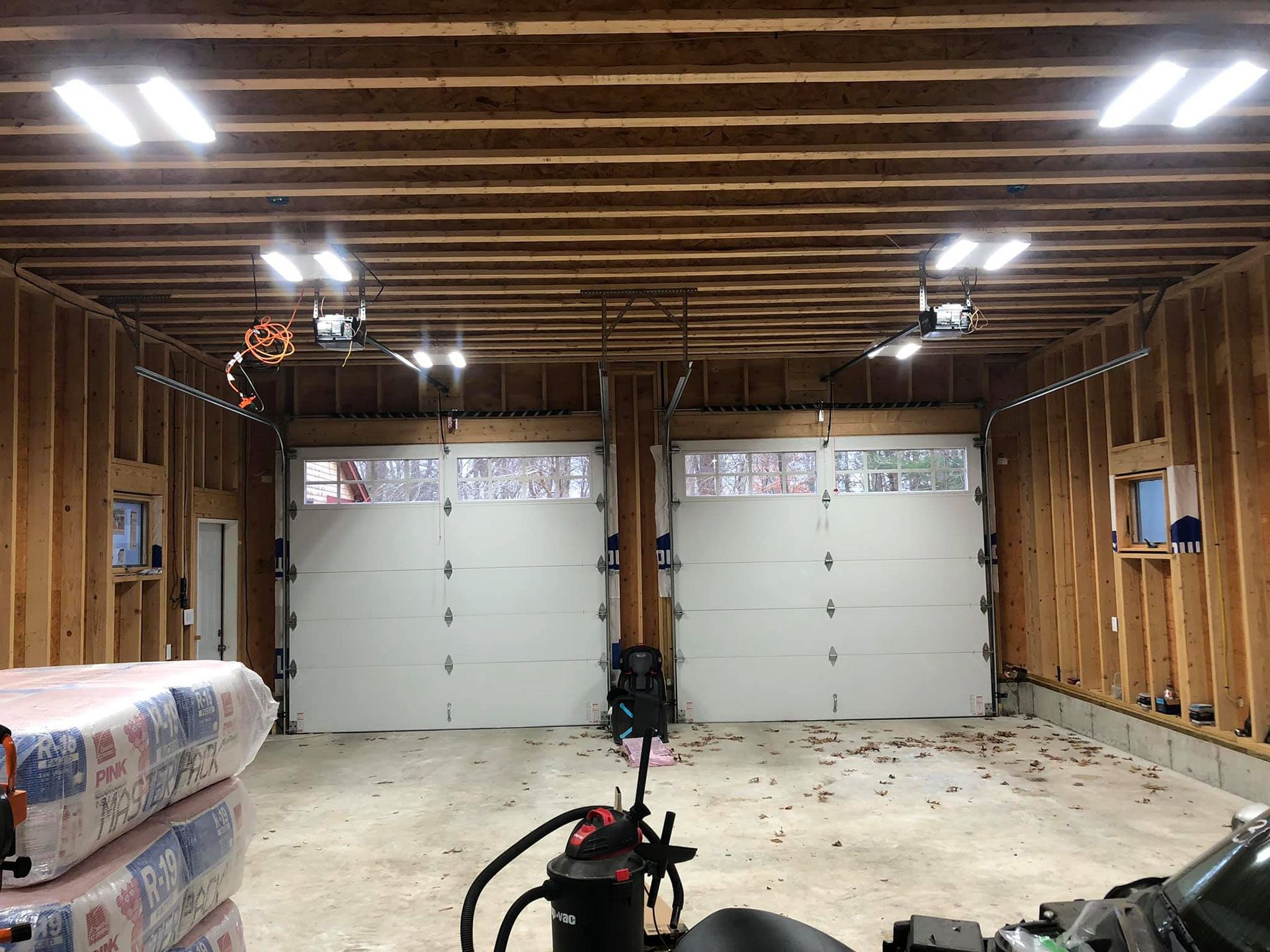 New garage interior with two white garage doors, exposed wood framing, and overhead lighting.