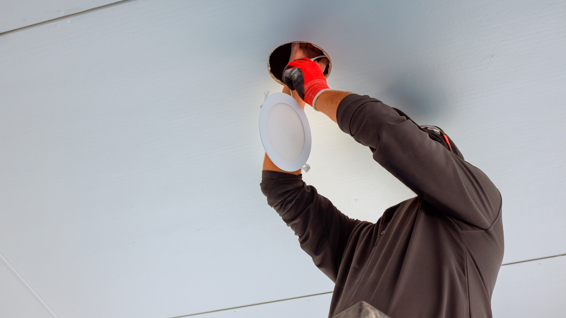 Person in red gloves installing a circular light fixture in a white ceiling.