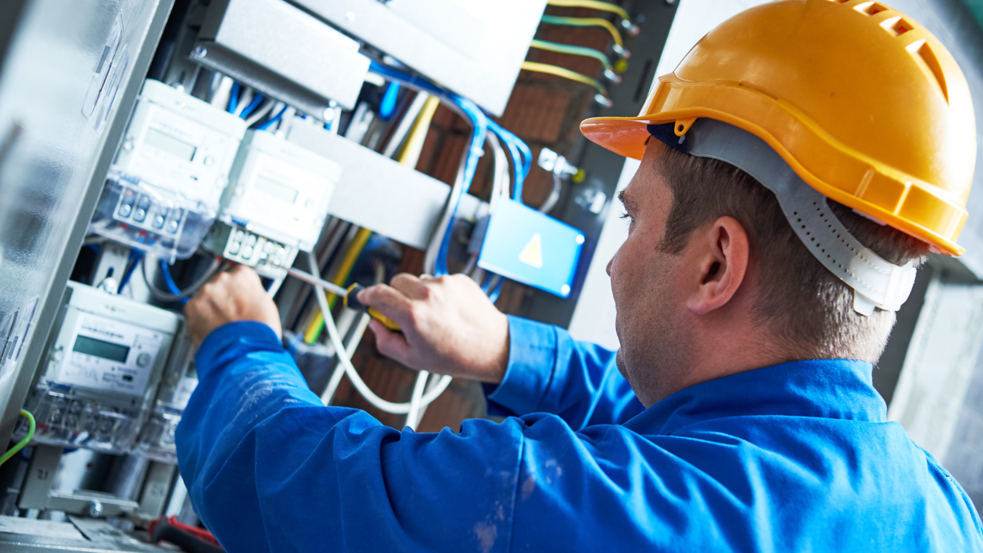 Electrician in yellow hard hat working on electrical panel, using screwdriver.