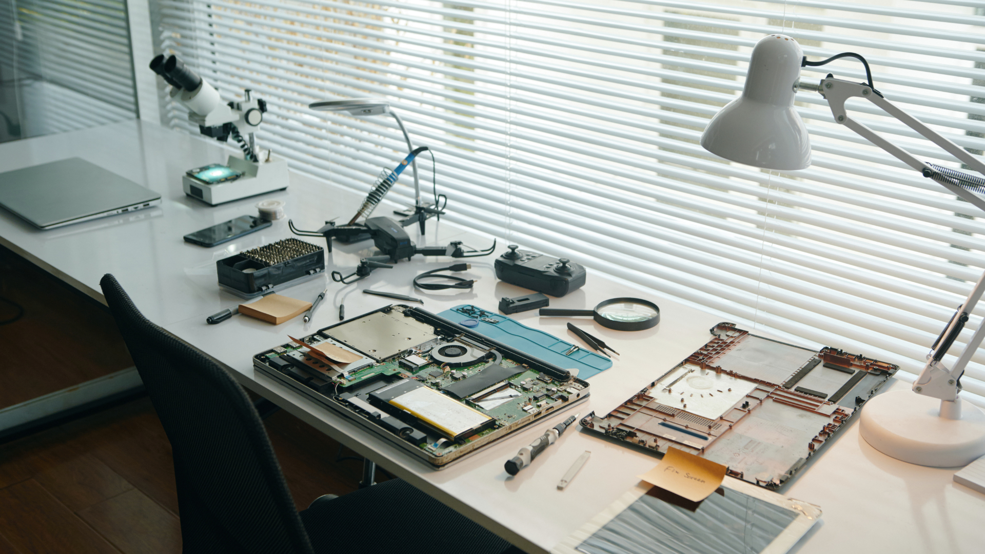 Laptop components and repair tools on a white desk by a window; microscope, lamp.