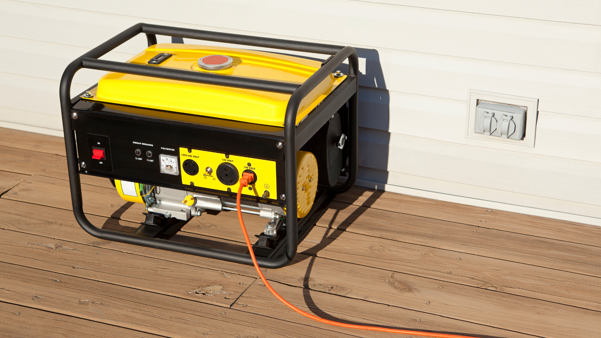 Yellow generator with black frame, orange cord plugged into it, on a wooden deck next to a white wall.