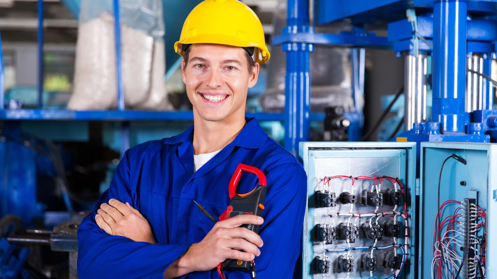 Smiling electrician in blue uniform, hard hat, holding a multimeter in an industrial setting.