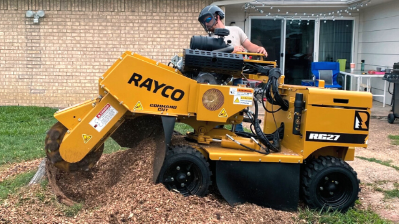 A person operates a yellow Rayco stump grinder, shredding a tree stump into wood chips in a residential yard.