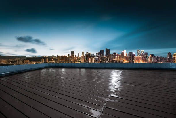 Rooftop view of a cityscape at dusk, reflecting lights on wet wooden deck; dark blue sky.