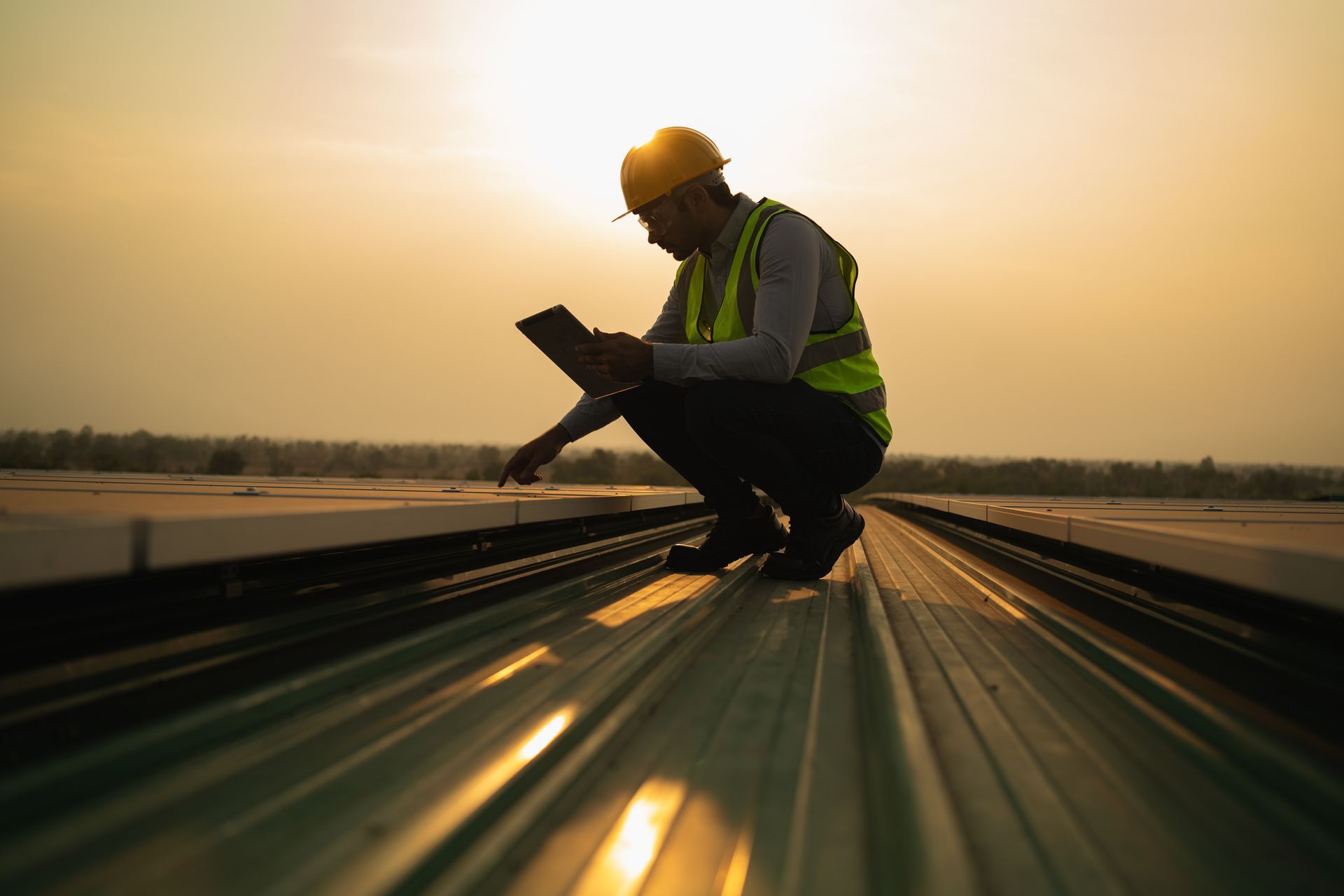 Construction worker on a rooftop, wearing a safety vest and helmet, inspecting with a tablet, sunset.