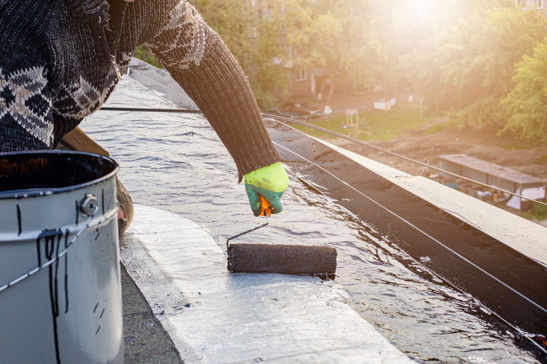 Person using a roller to apply black sealant on a flat roof, near a bucket of sealant.