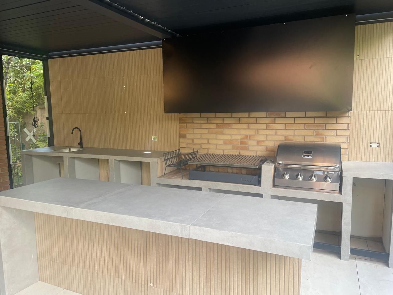 Cocina al aire libre con parrilla, fregadero y barra bajo un dosel negro; detalles de ladrillo y madera.