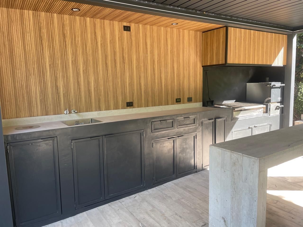 Cocina al aire libre con paneles de madera, gabinetes grises, encimeras de concreto y barra.