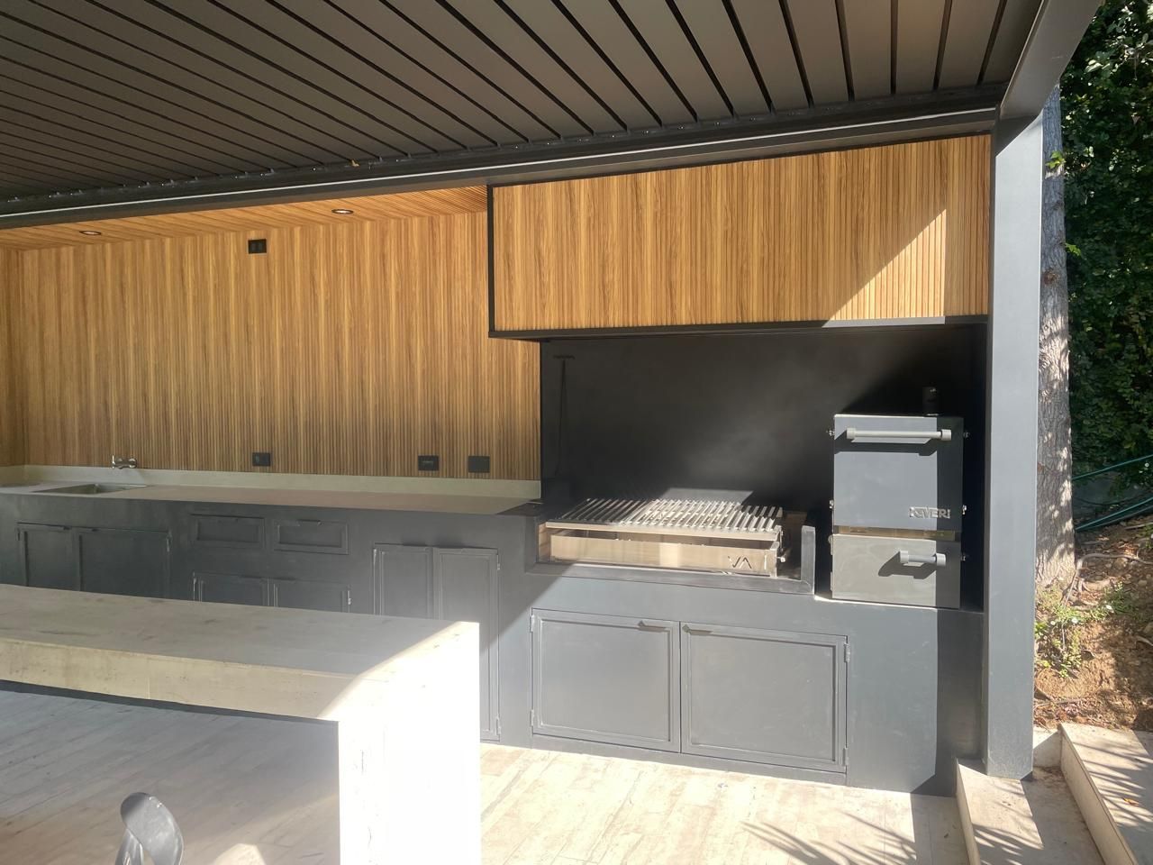 Cocina exterior con parrilla, paneles de madera y encimera de hormigón. Tonos de madera gris y natural.