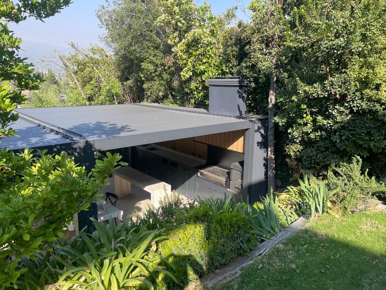 Zona de barbacoa exterior cubierta con techo gris y chimenea, rodeada de árboles y vegetación.