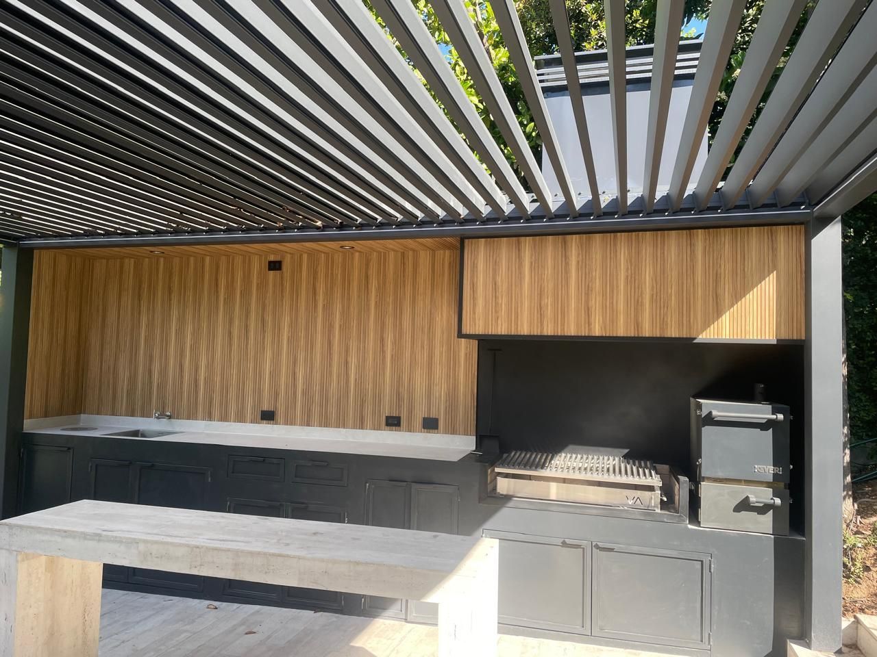 Cocina al aire libre con gabinetes grises, paneles de madera y una encimera de concreto bajo un techo de listones.