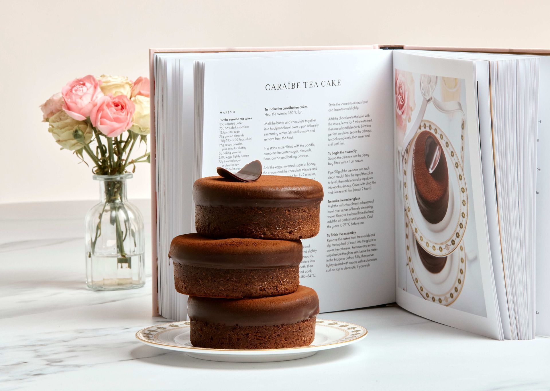 Stack of chocolate cakes, open cookbook, vase of flowers on white surface.