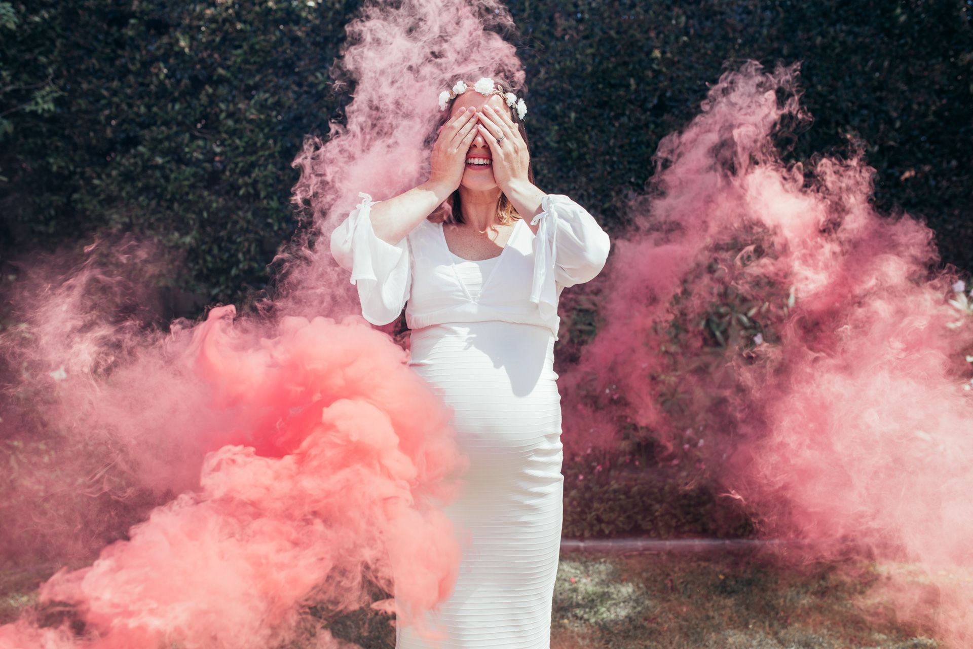 A pregnant woman in a white dress is standing in front of pink smoke.