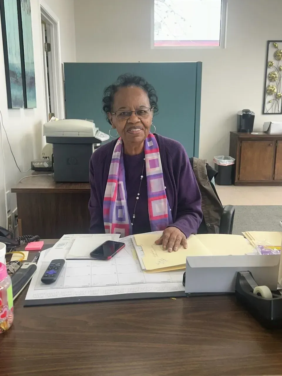 Woman wearing purple cardigan and scarf sits at desk, smiling.