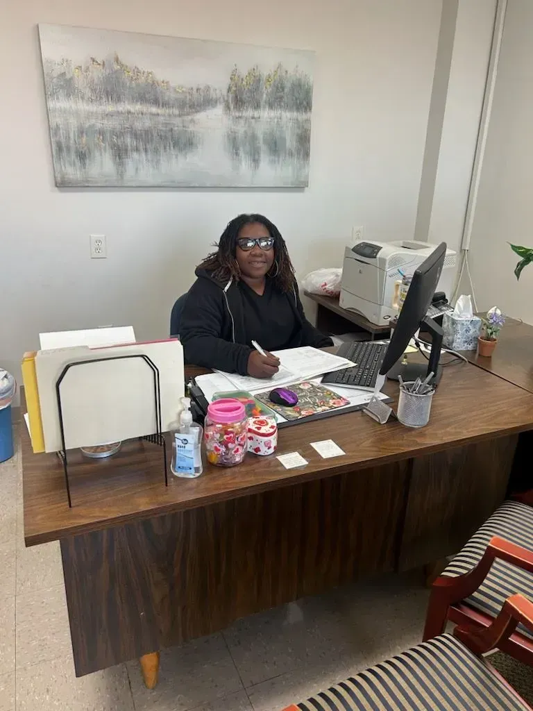 Person sitting at a desk, smiling. Desk has paperwork, computer, and candy. Art on wall.
