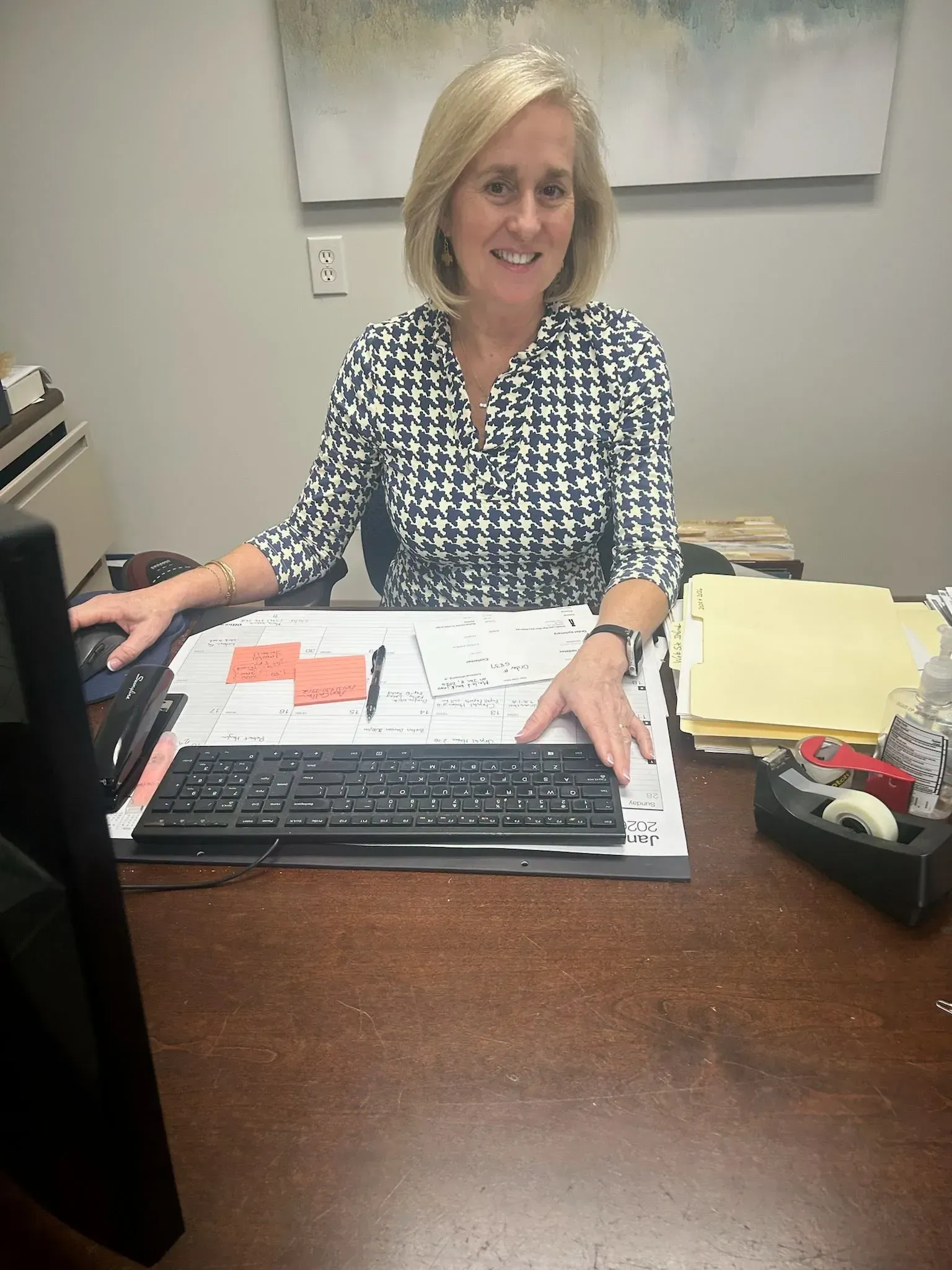 Woman at a desk with a keyboard, papers, and a computer monitor, smiling at the camera.