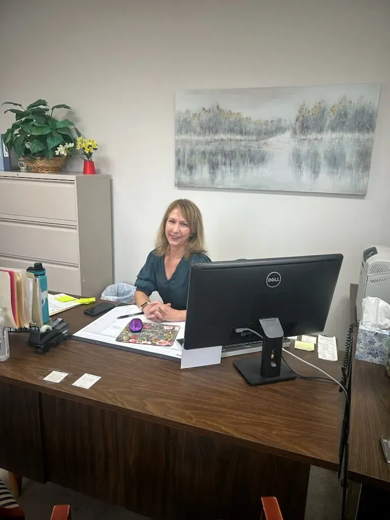 Woman sitting at a desk with a computer, smiling. Behind her, a painting and a cabinet.