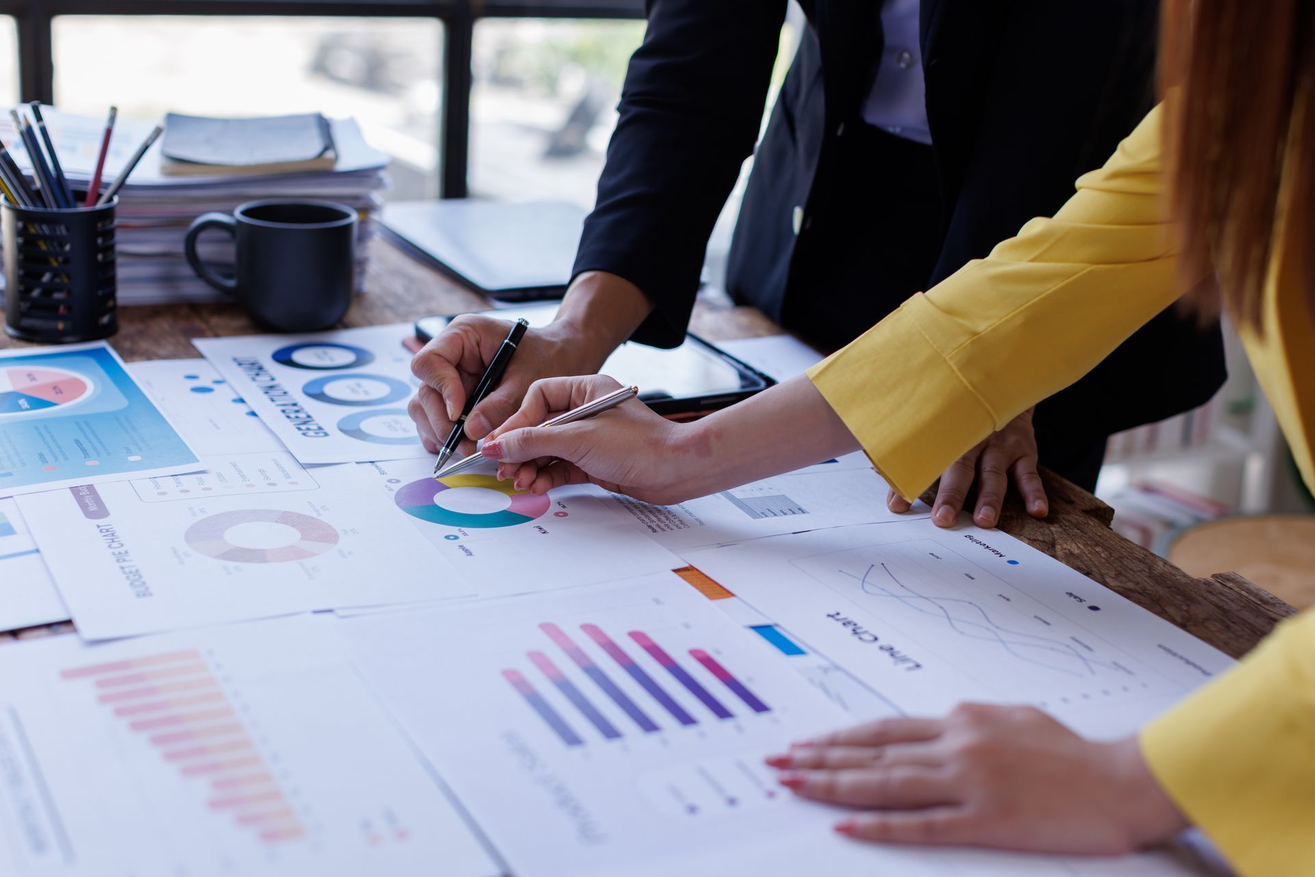 Two people analyzing charts and graphs on a desk with a laptop and coffee mug.
