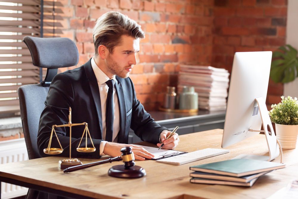 Lawyer in a suit working on a computer, writing at a desk, scales of justice and gavel nearby.