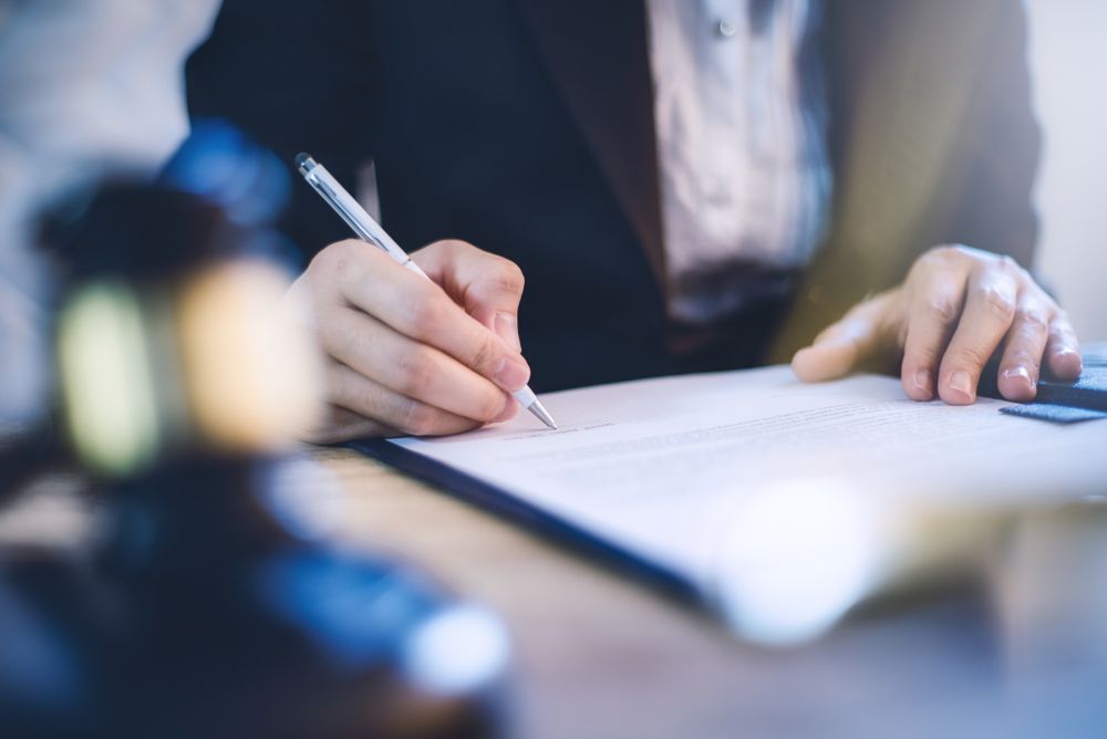 Person in suit writing on paper with a pen; gavel in the foreground.