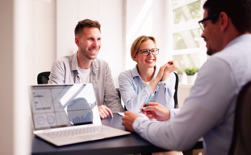 Couple smiling, meeting with advisor at a desk with a laptop, bright room.