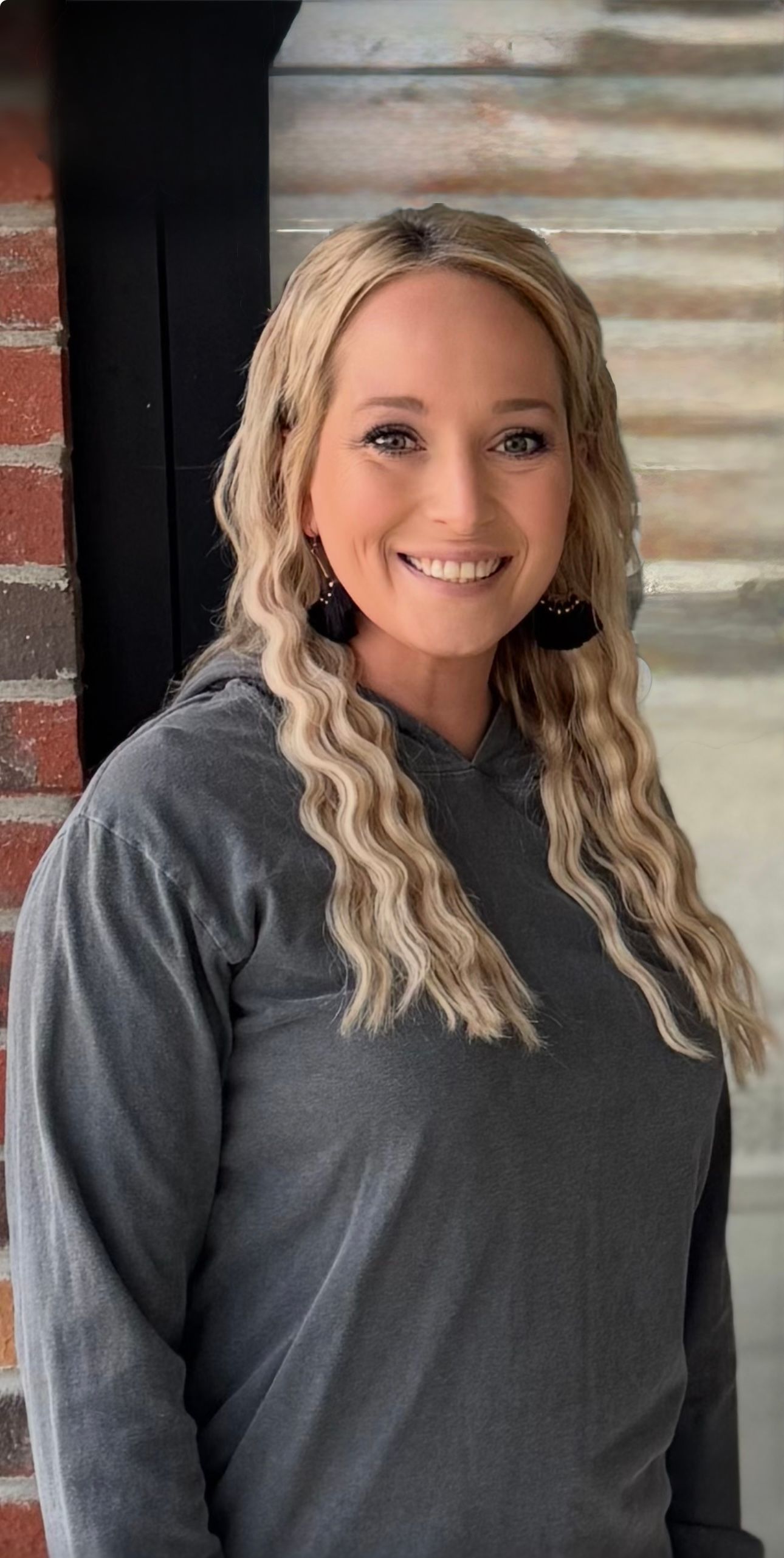 Woman with curly blonde hair smiles, wearing a gray sweatshirt and black earrings, against a brick wall.