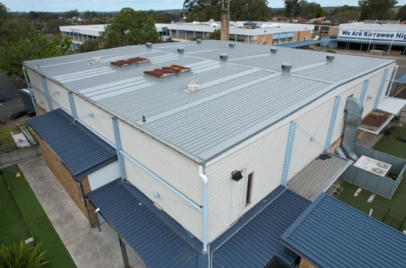 A rectangular, light gray building with a corrugated metal roof. A smaller structure with a blue roof is in front.
