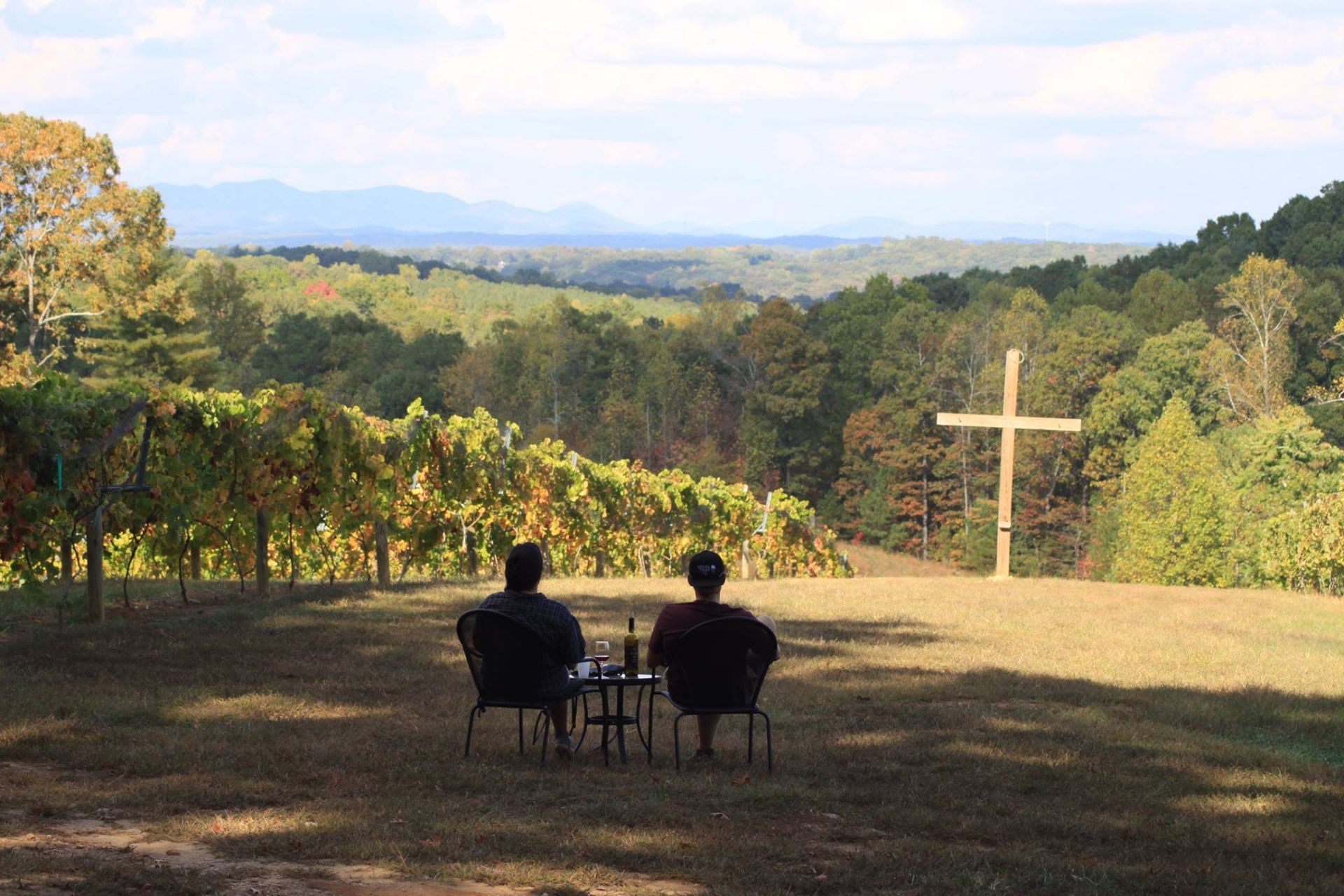 Two people sit in a field with a cross in the background