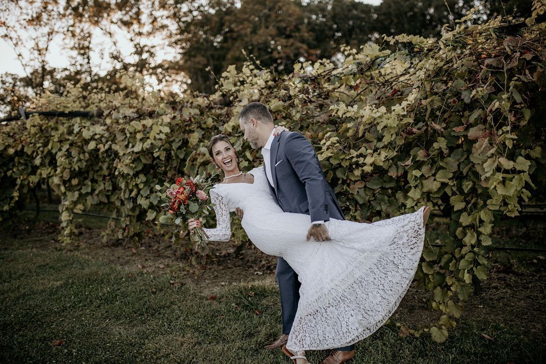 A man is carrying a bride in his arms in a vineyard.