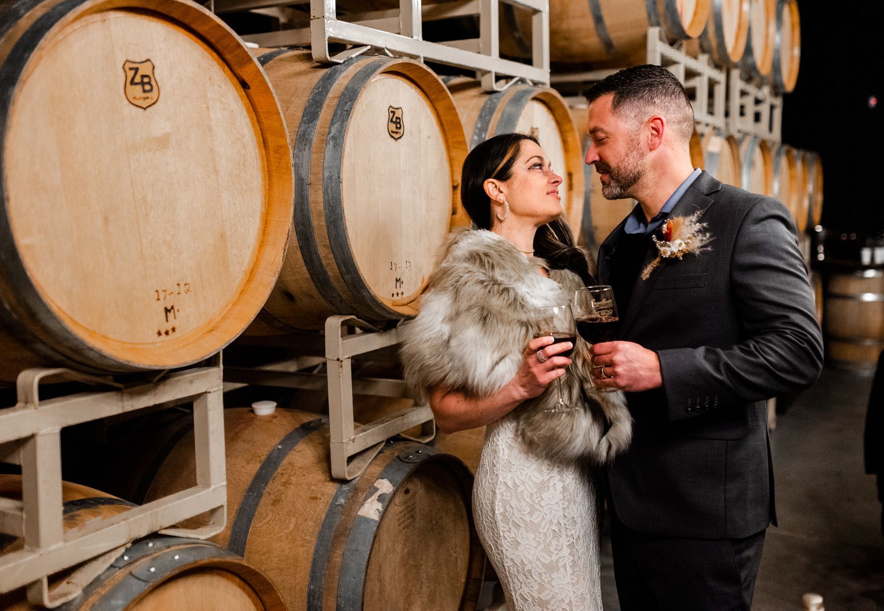 A bride and groom are standing next to wine barrels in a wine cellar.