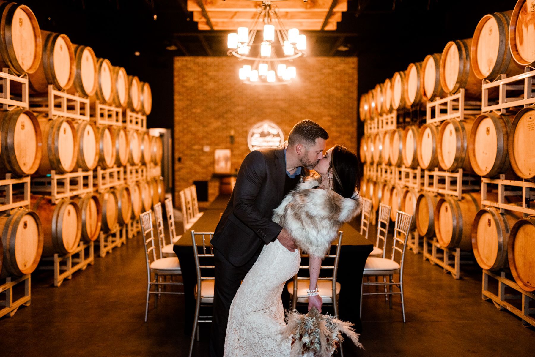A bride and groom are kissing in a wine cellar filled with wine barrels.