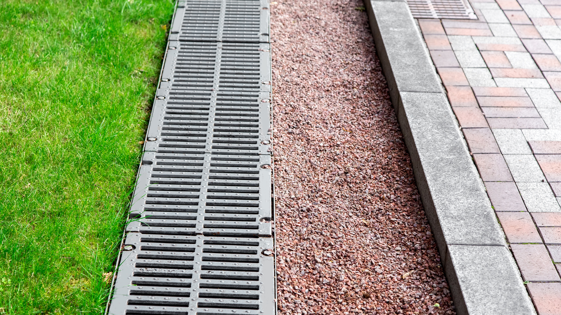 A drainage system with a metal grate separates green grass from red gravel and brick paving.