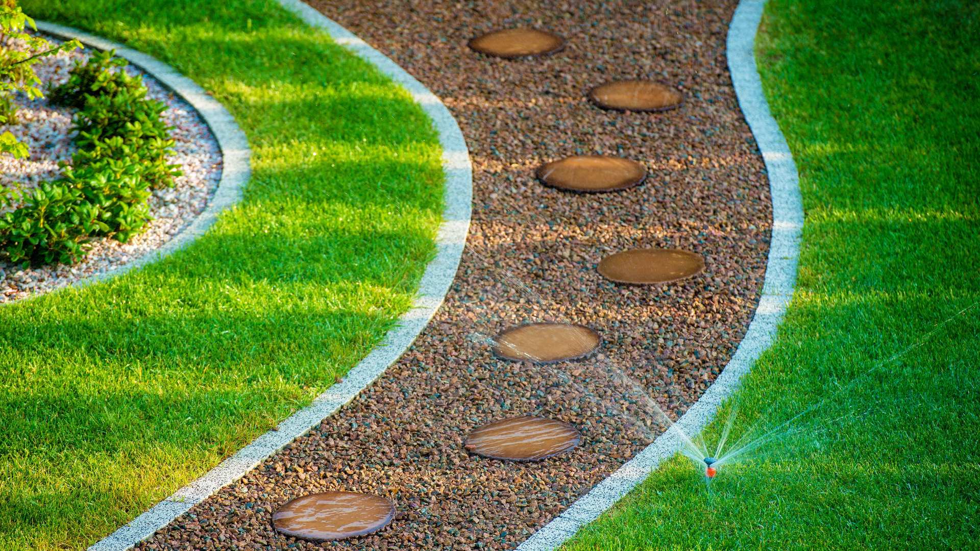 Curving stone path with stepping stones through brown gravel, bordered by green grass and white stones.
