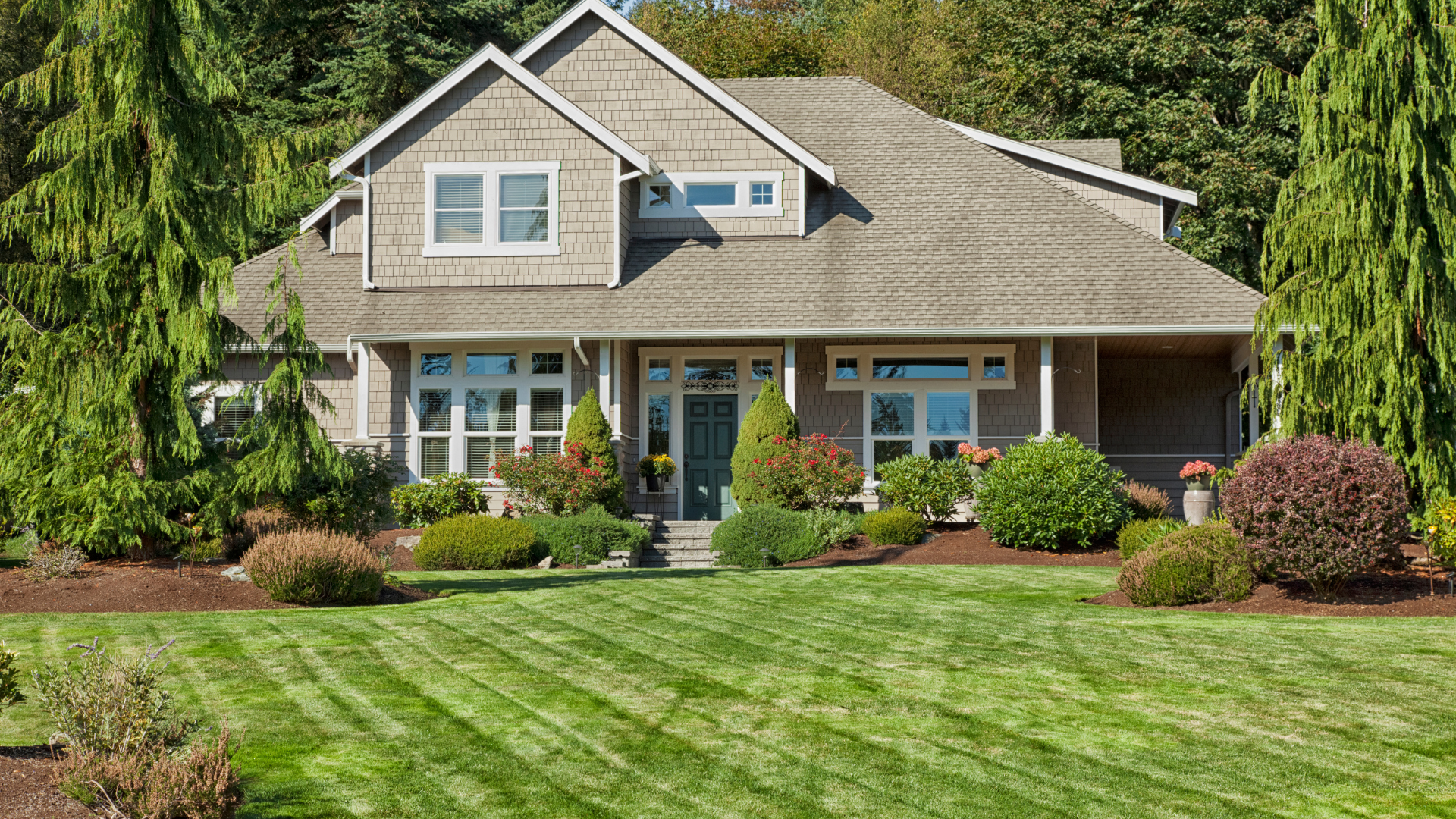House with light brown siding, green lawn, trees, and shrubs.
