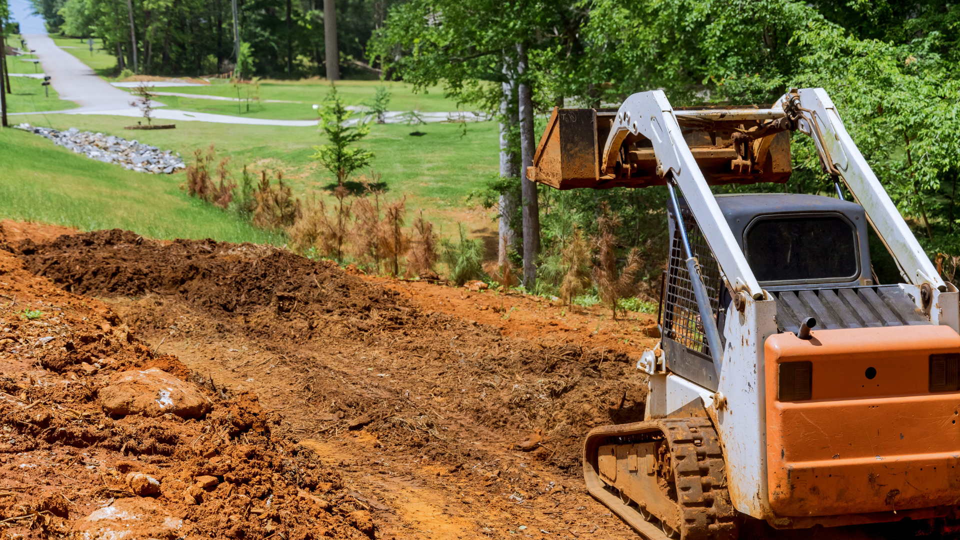 Skid steer loader on a hillside, moving dirt. Orange, white, and black machinery against a green landscape.