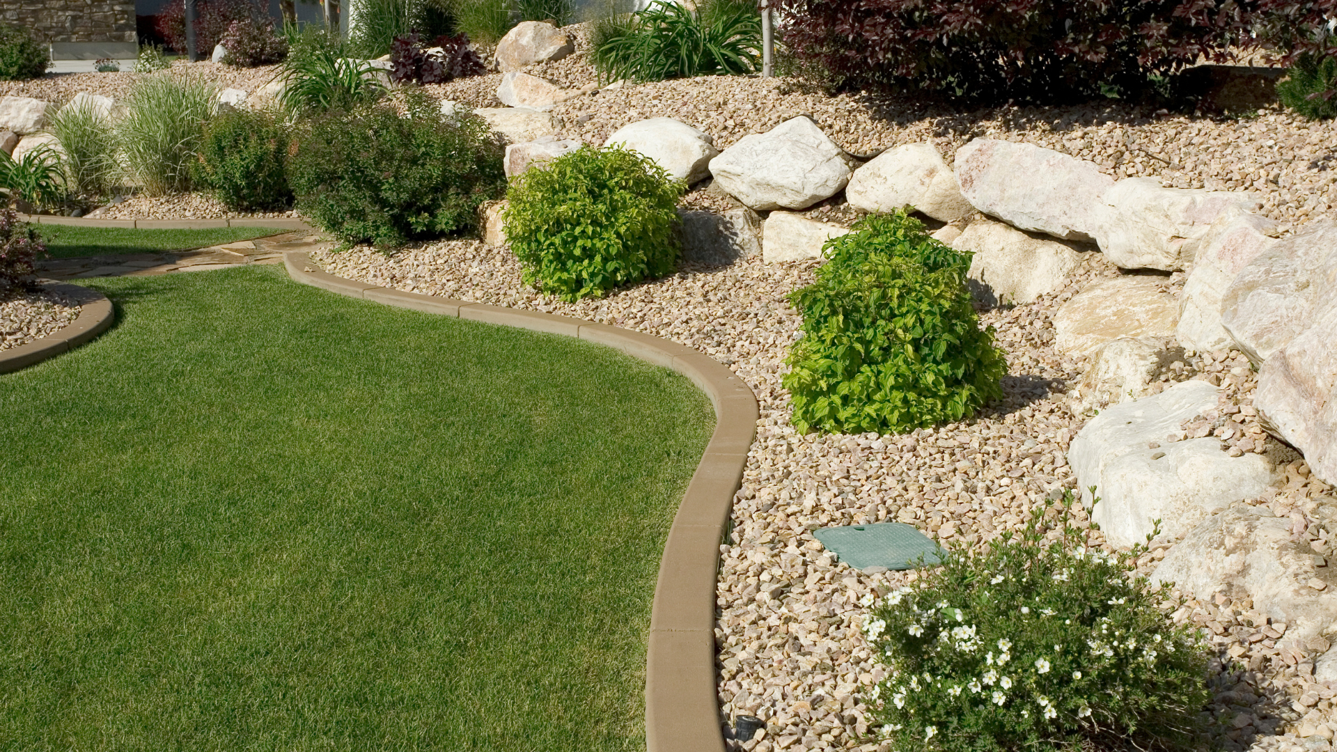 Lush green lawn borders a bed of tan gravel and large rocks with small green shrubs and bushes.