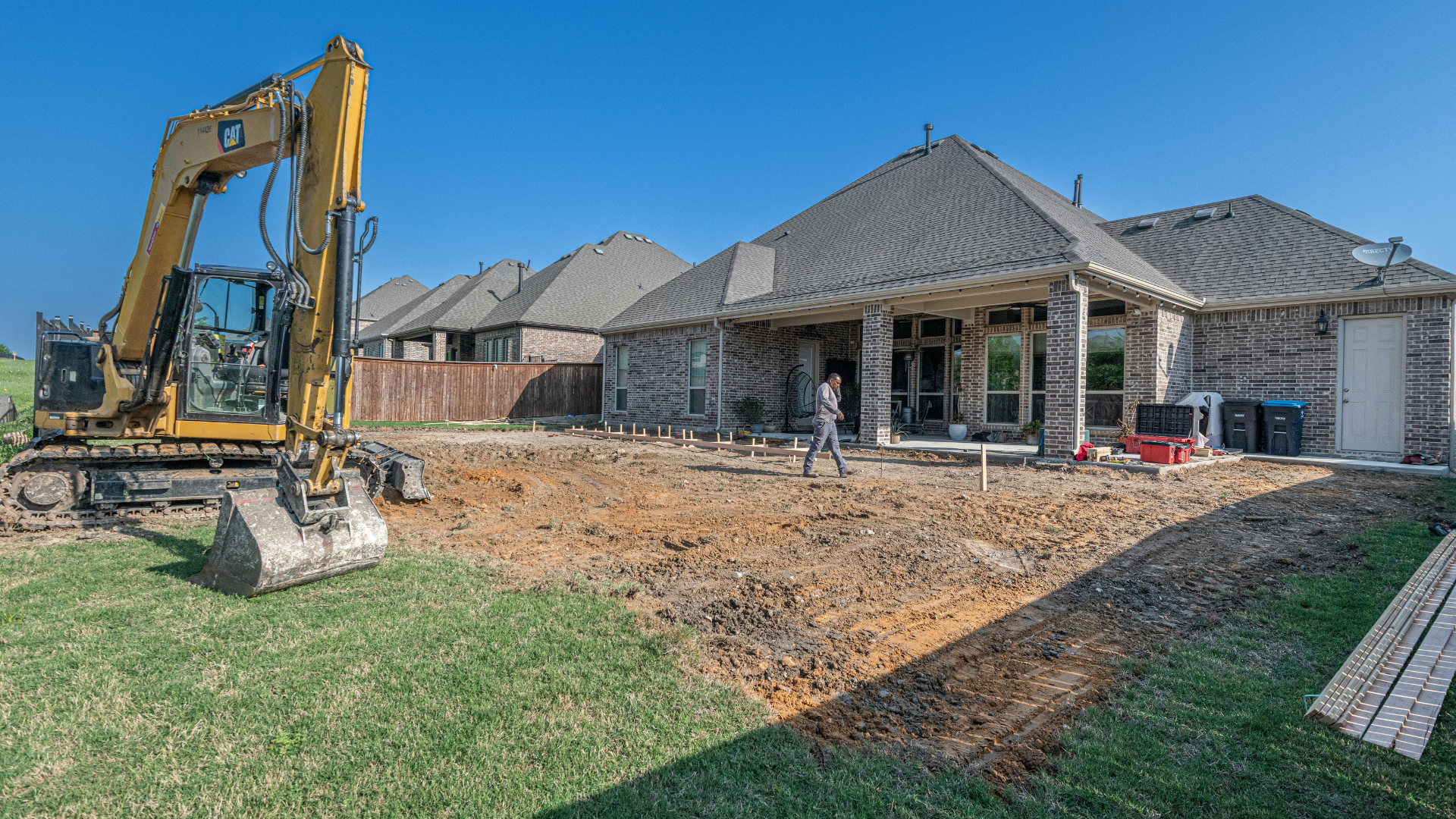 Excavator digging in a backyard, with a house in the background and a person standing nearby.