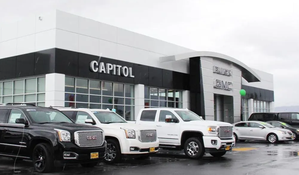 Capitol Buick GMC dealership with vehicles on display. White and black building, overcast sky.
