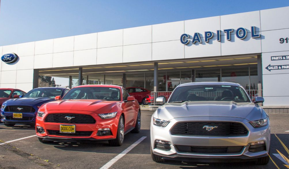 Ford Mustang cars parked in front of Capitol Ford dealership, under blue sky.