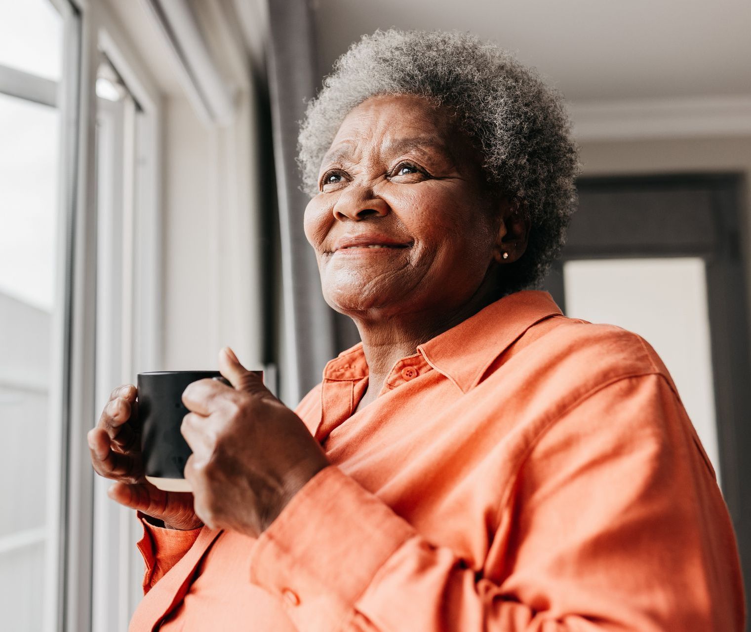 An elderly woman is holding a cup of coffee and looking out the window.