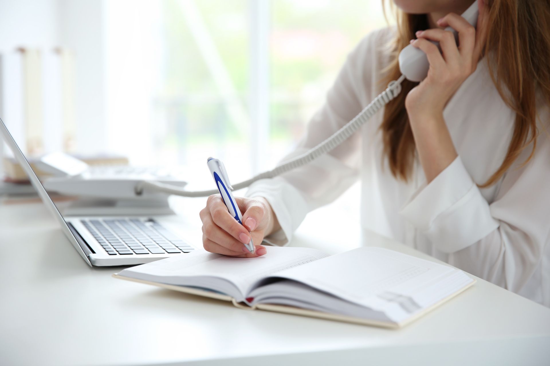 Woman writing in a notebook while holding a landline phone to her ear; laptop nearby on a white desk. Consulting services in Halfmoon NY
