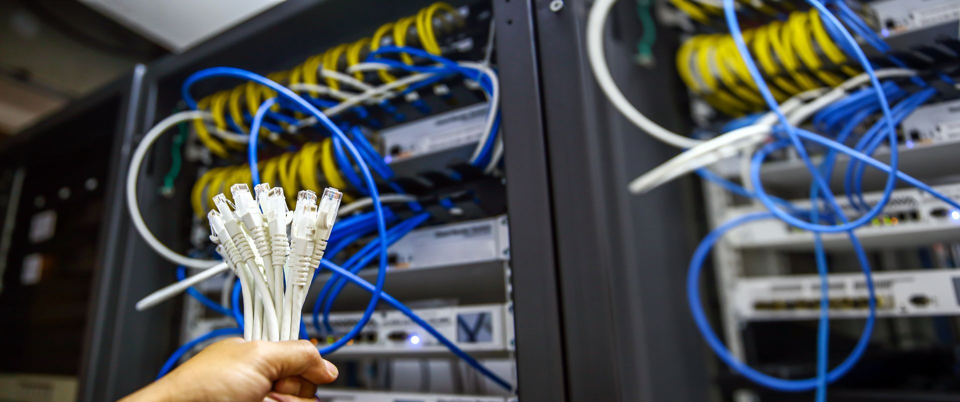 Hand holding a bundle of white network cables near a server rack with blue and yellow cables. Core cabling in Halfmoon NY