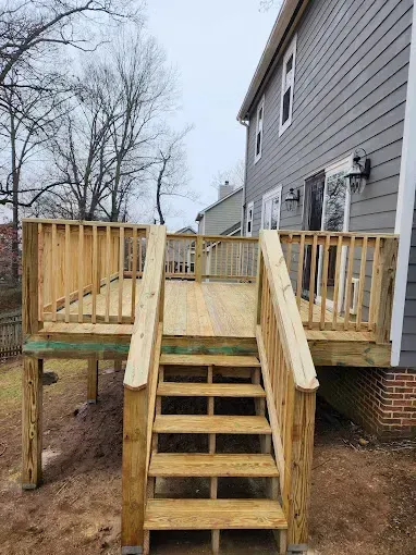 Wooden deck with stairs, attached to a gray house. Natural wood color. Cloudy sky.