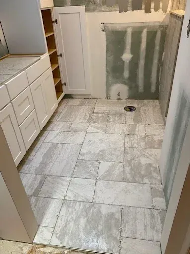 Bathroom under construction: white cabinets, gray tile floor, unfinished walls, and a drain.